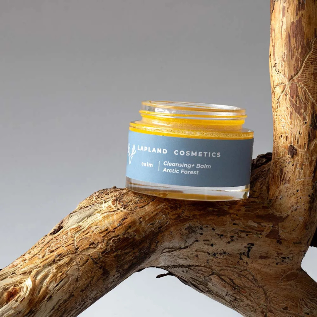 Open jar of Lapland Cosmetics Cleansing+ Balm Arctic Forest resting on textured driftwood against a plain gray background.